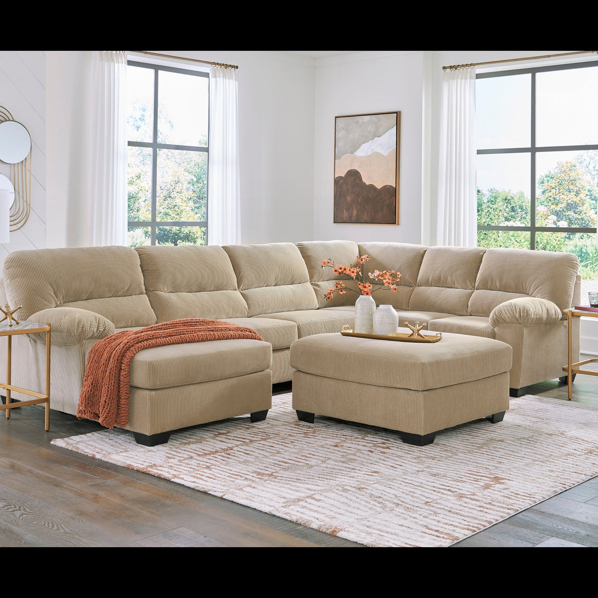 A beige fabric sectional sofa with a matching chaise ottoman in a bright, modern living room. The sectional is styled with an orange throw blanket and a small decorative tray on top, set on a patterned area rug with side tables and lamps.