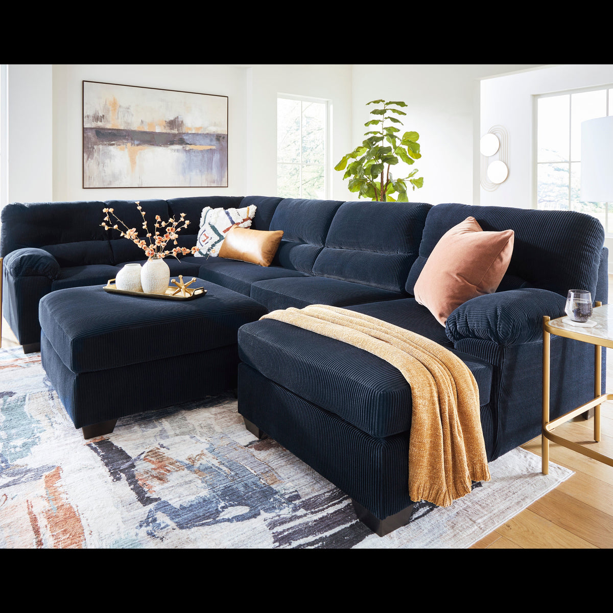 A large sectional sofa in dark navy blue corduroy fabric, styled in a bright, modern living room with a matching ottoman, decorative pillows, a throw blanket, and a patterned area rug.