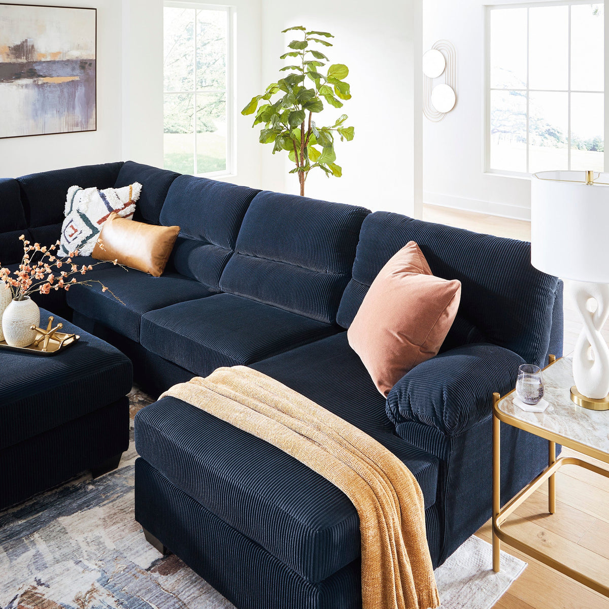 A dark navy blue sectional sofa with a matching ottoman in a bright, modern living room. The sofa is styled with a peach throw pillow, a tan throw blanket, and decorative pillows, set on a patterned rug with light wood flooring and ceiling beams.