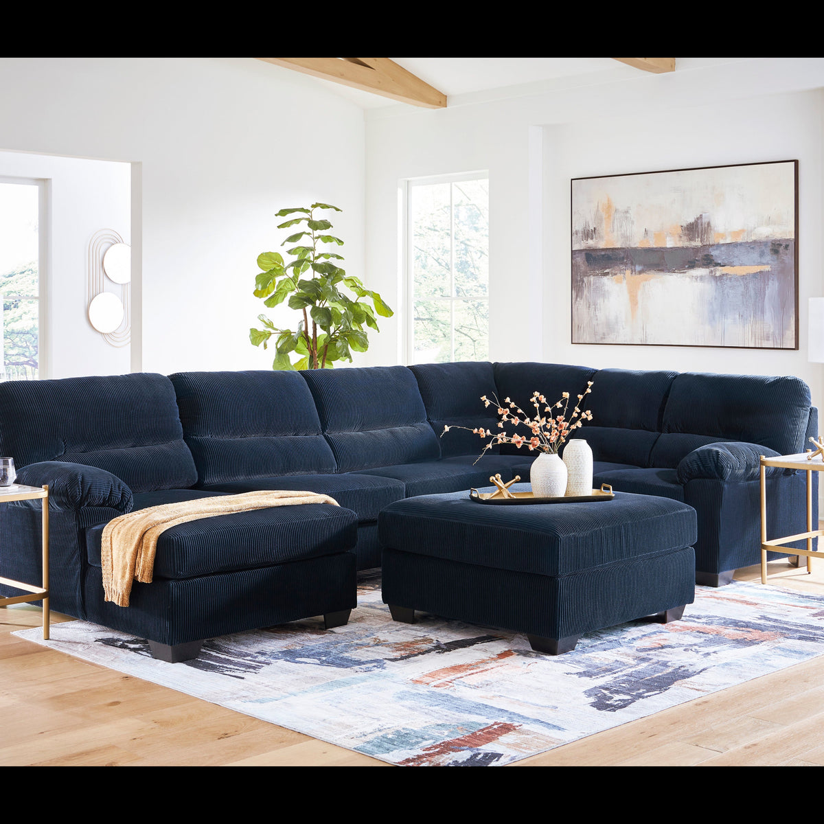 A dark navy blue sectional sofa with a matching ottoman in a bright, modern living room. The sectional is styled with a gold-framed side table, white table lamps, a patterned area rug, and a green potted plant in the background.
