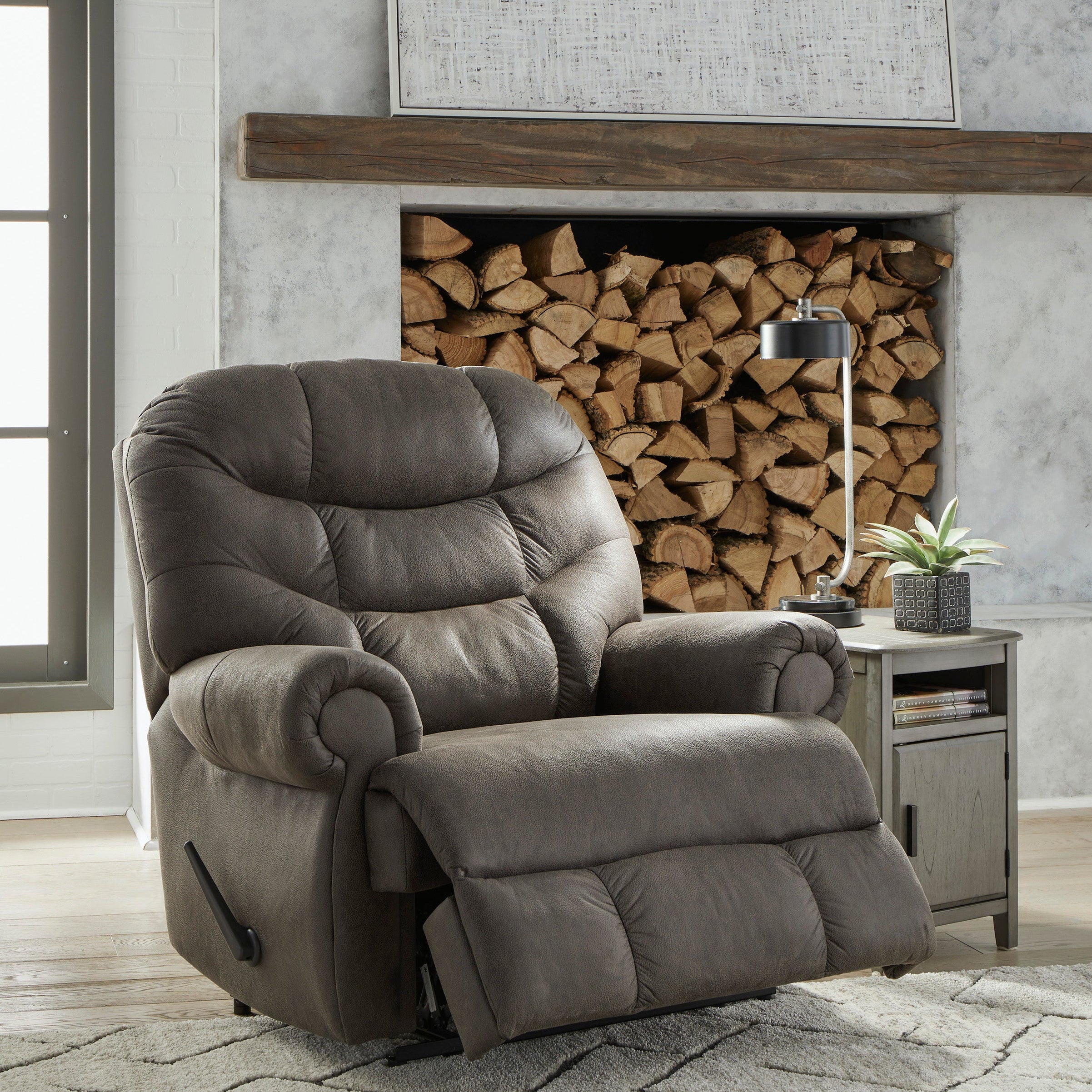 Gray recliner chair in a living room setting with a fireplace and wooden logs.