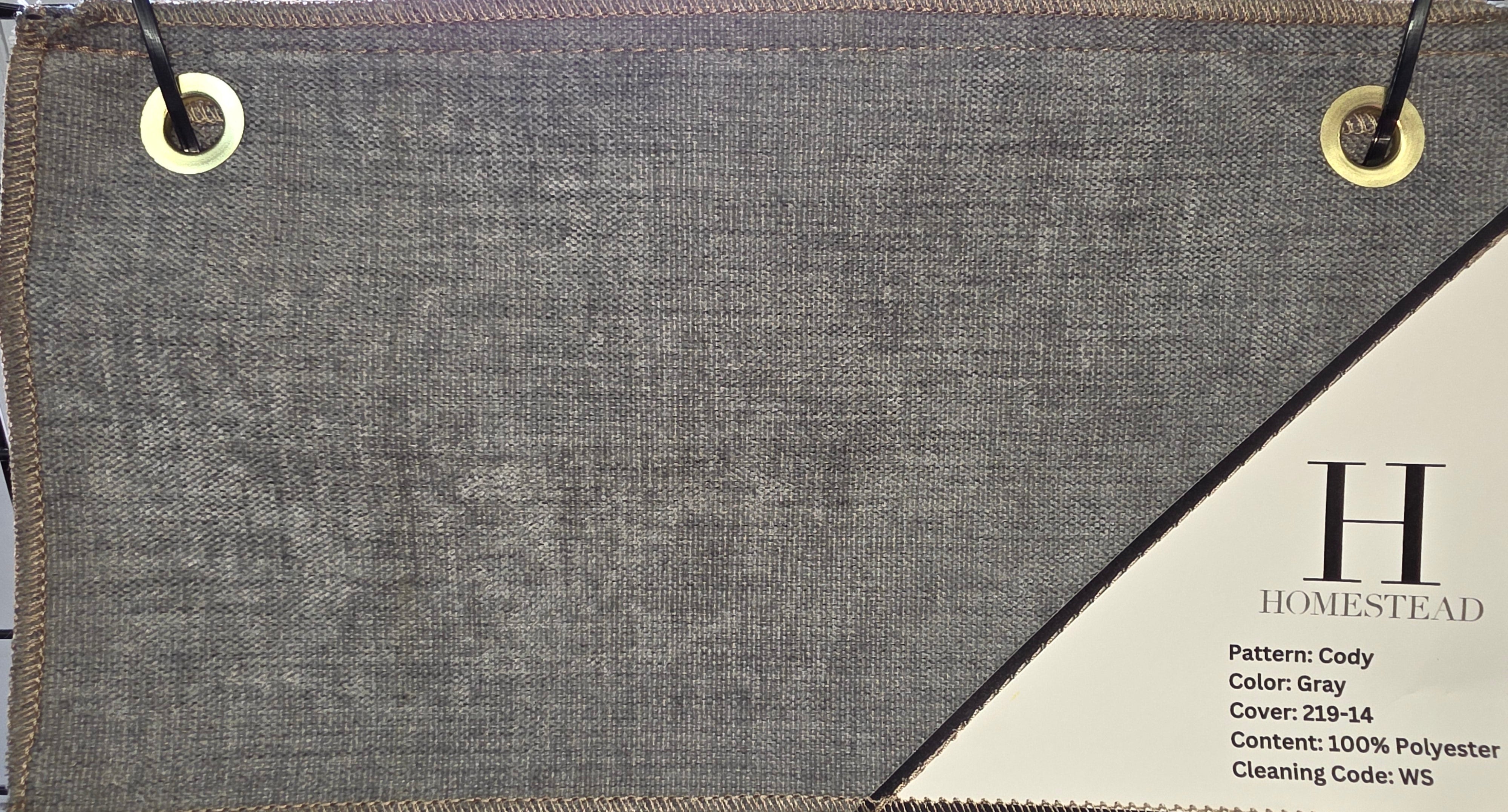 A fabric sample swatch labeled H Homestead Pattern: Cody, Color: Gray, displayed on a rack. The swatch shows a textured woven gray material with a diagonal white printed section containing product details, and brass grommets are visible at the corners.