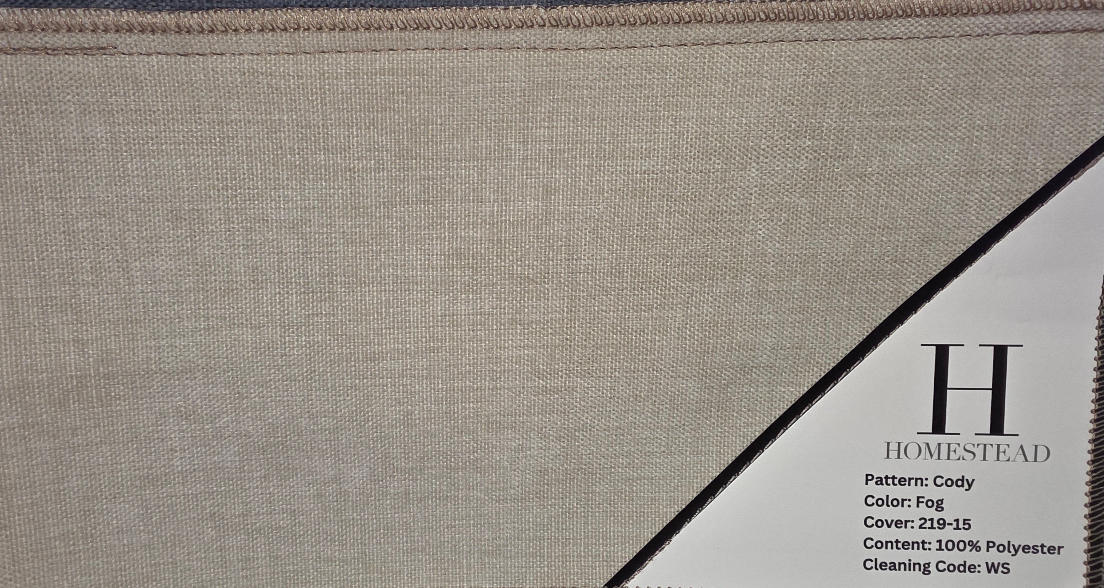A fabric swatch sample labeled H Homestead Cody, showing a light beige woven texture with a diagonal black label overlay and additional fabric samples below on a wire display.