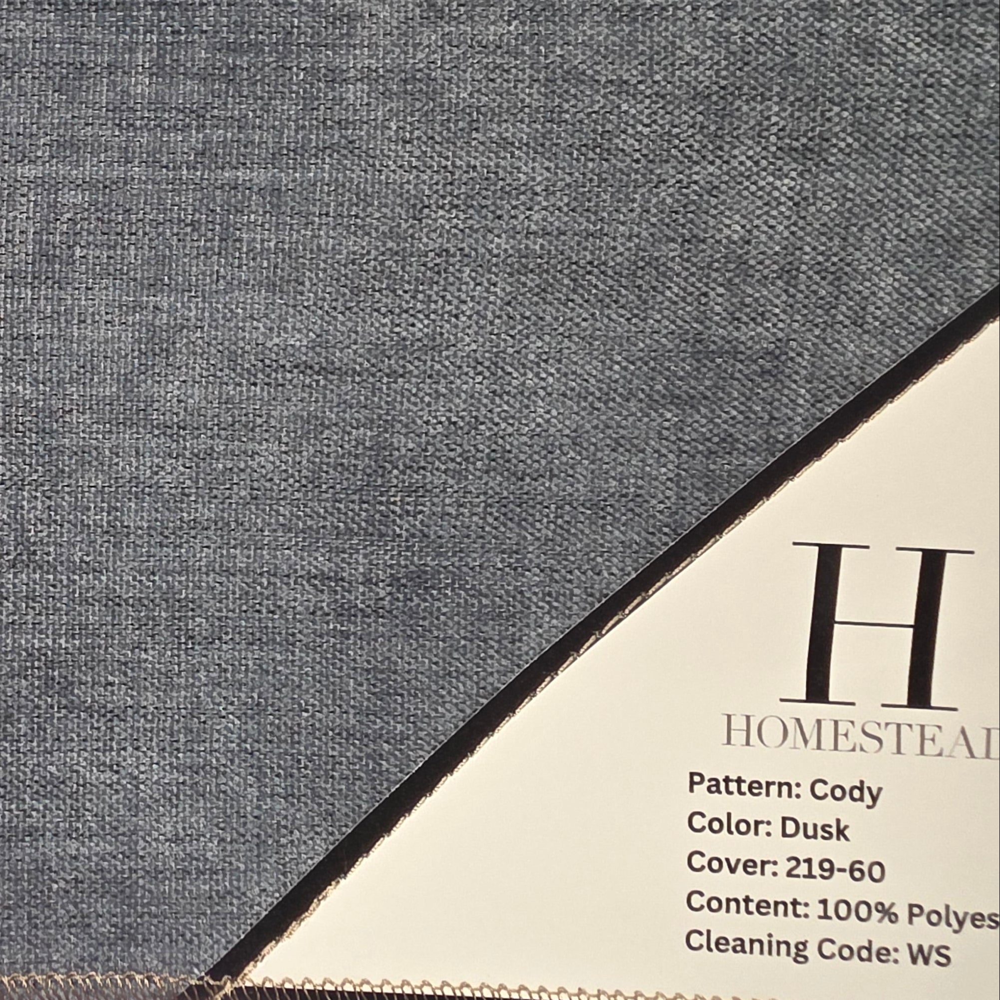 A close-up of a dark gray textured outdoor canopy fabric with a beige corner label showing product details for 'Homesteal' in the 'Cody' pattern.