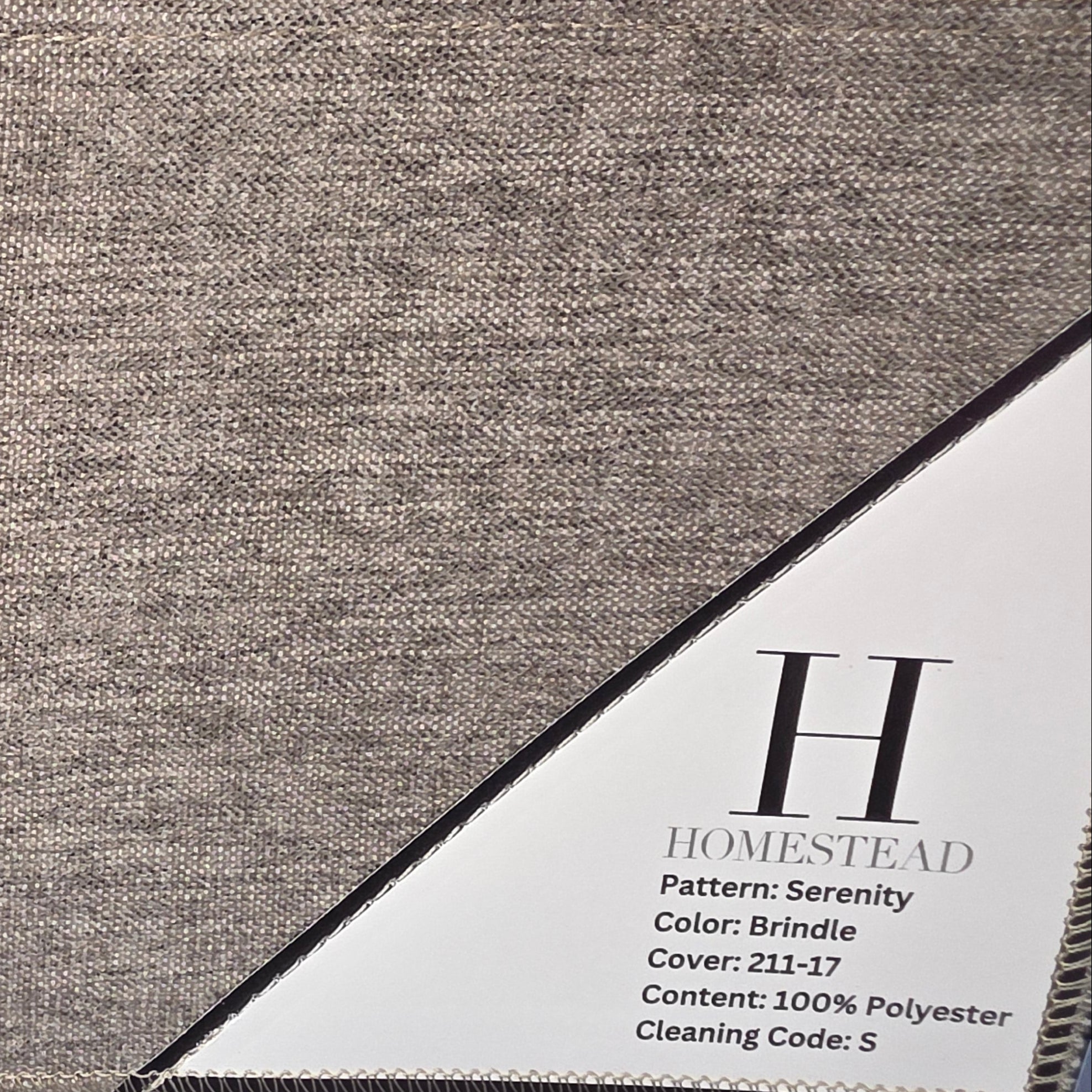 A close-up of a woven fabric swatch labeled Homestead, showing a heathered taupe-beige textile with a white paper tag displaying product details.
