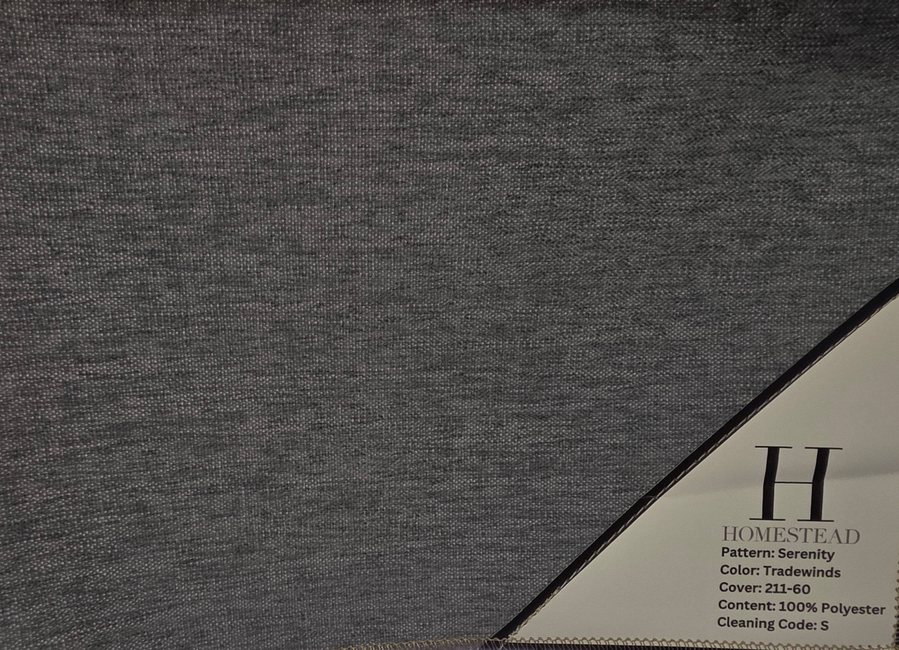 A close-up of a gray woven fabric sample hanging from a black wire rack, with a printed corner label reading Homestead and listing details. A bright window is visible in the background behind the rack.