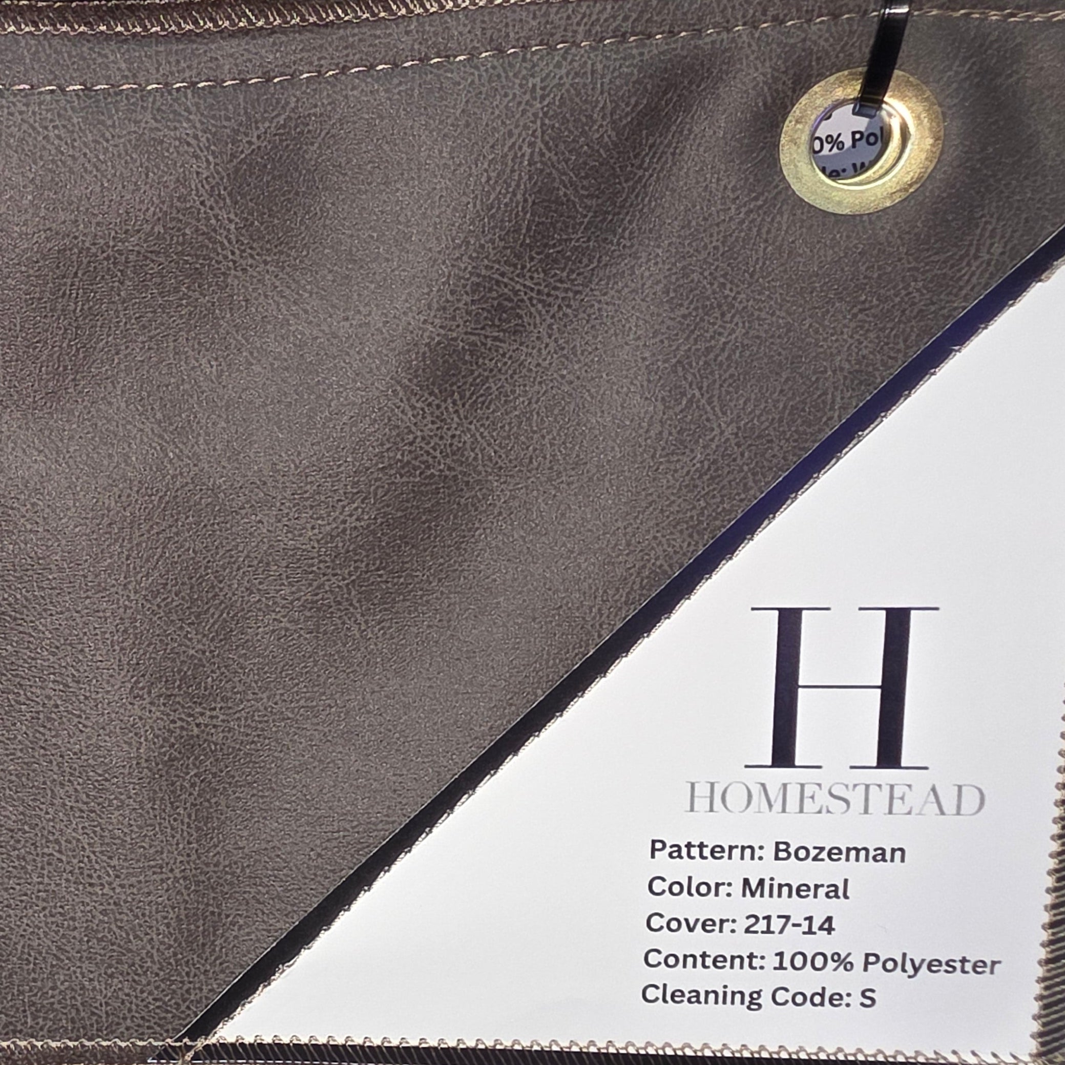 A close-up of a Homestead clutch or envelope-style handbag in a taupe-gray mineral finish, with a white printed product tag attached and a brown suede-like section visible along the bottom edge.
