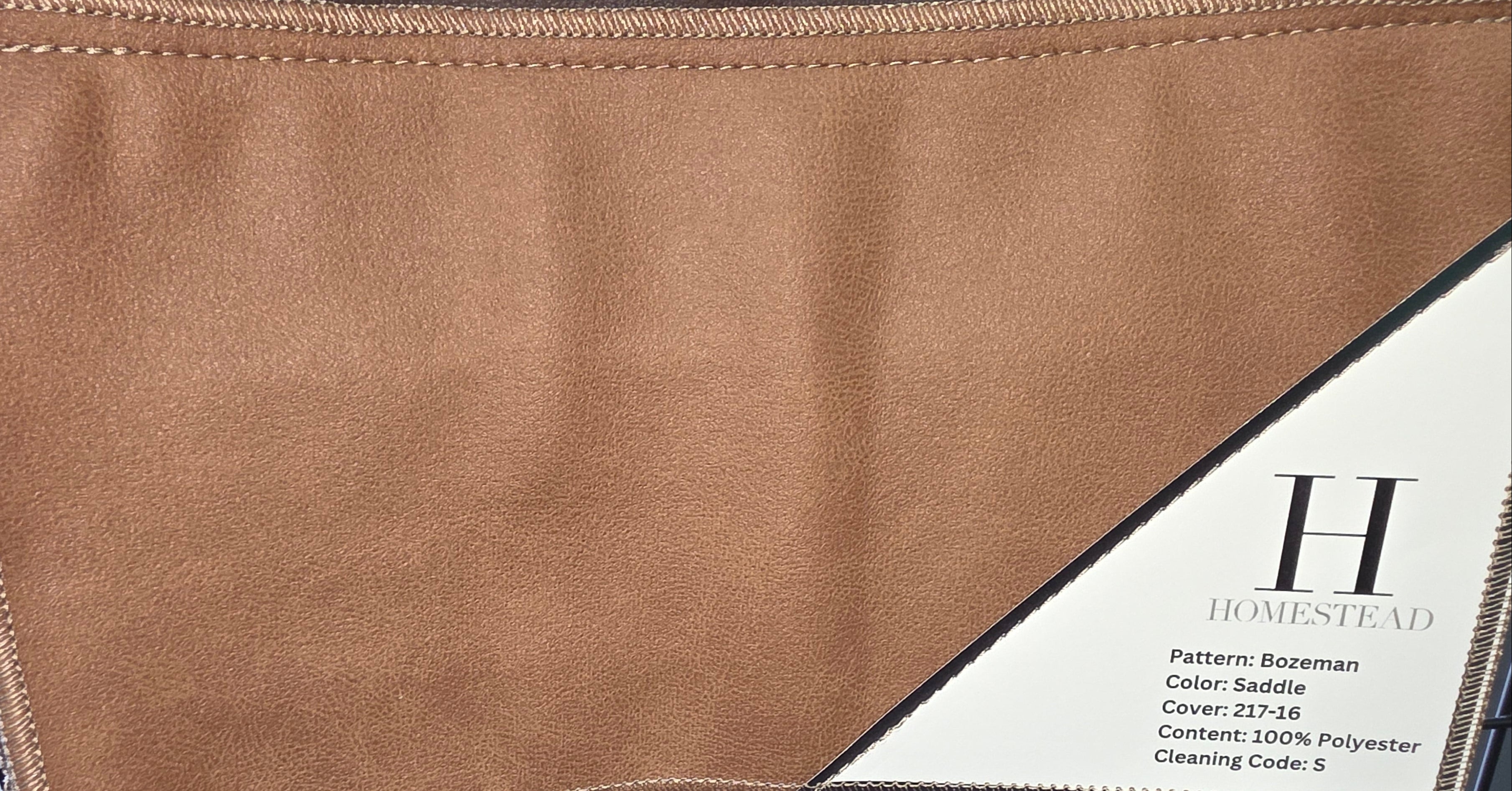A close-up of a brown textured fabric or leather material sample with visible stitching and a printed label reading H Homestead, showing color and pattern details.