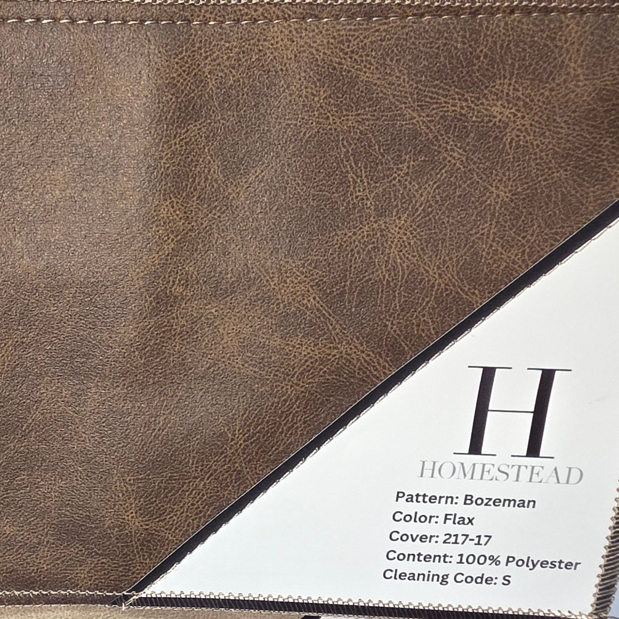 A close-up of a brown textured fabric or faux-leather material sample with stitched edges, shown against a white grid background. A printed label card for Homestead is partially visible on the lower right, showing product details.