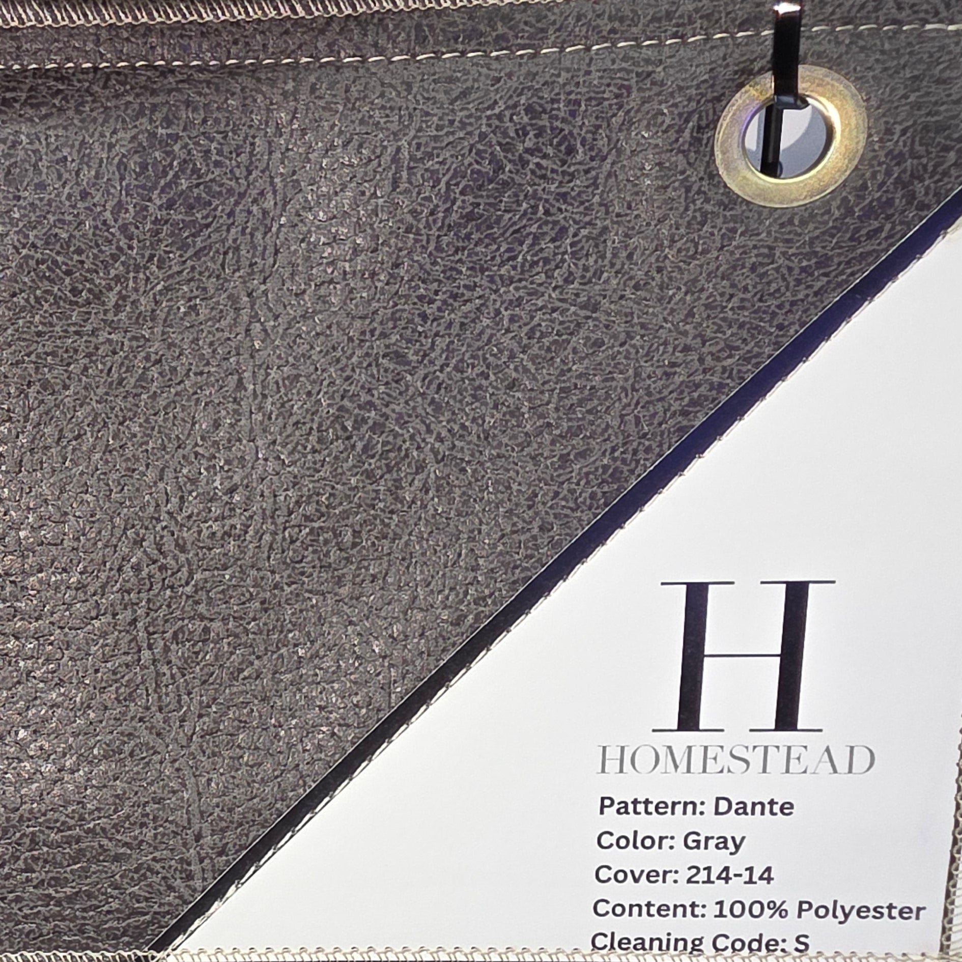 Close-up of a hanging fabric or faux-leather panel sample with a pebbled texture, showing a gray upper section and a darker brown lower section. A white printed label on the right reads 'H HOMESTEAD' with product details.