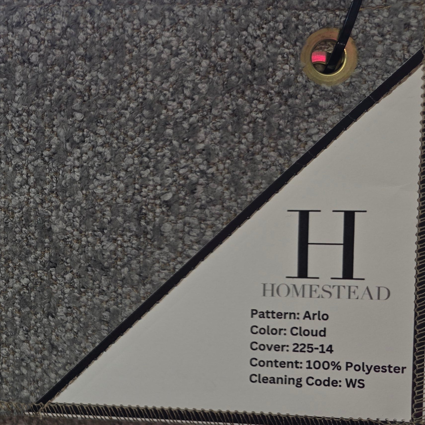 A close-up of a carpet sample hanging on a display rack. The sample has a textured, speckled cloud-like pattern in gray and beige tones, with a white paper label attached that reads 'H HOMESTEAD' and includes product details.