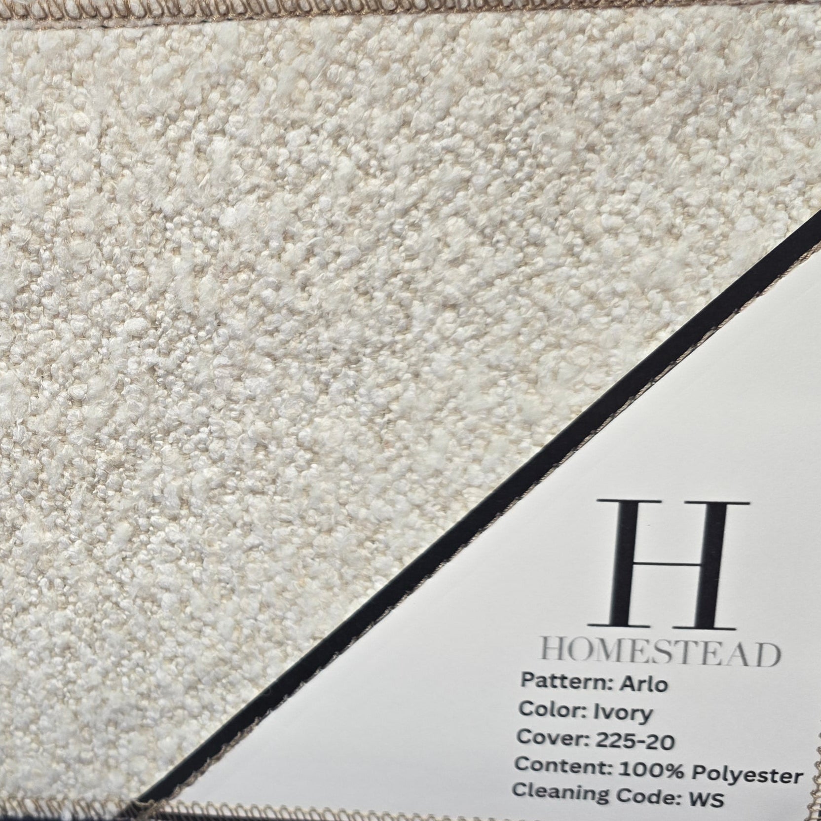 A close-up of a textured area rug sample hanging on a display rack. The top visible rug section is a dark navy blue with a subtle floral pattern, and a large cream/beige rug sample is shown in front with a printed label reading Homestead and Pattern Aids.