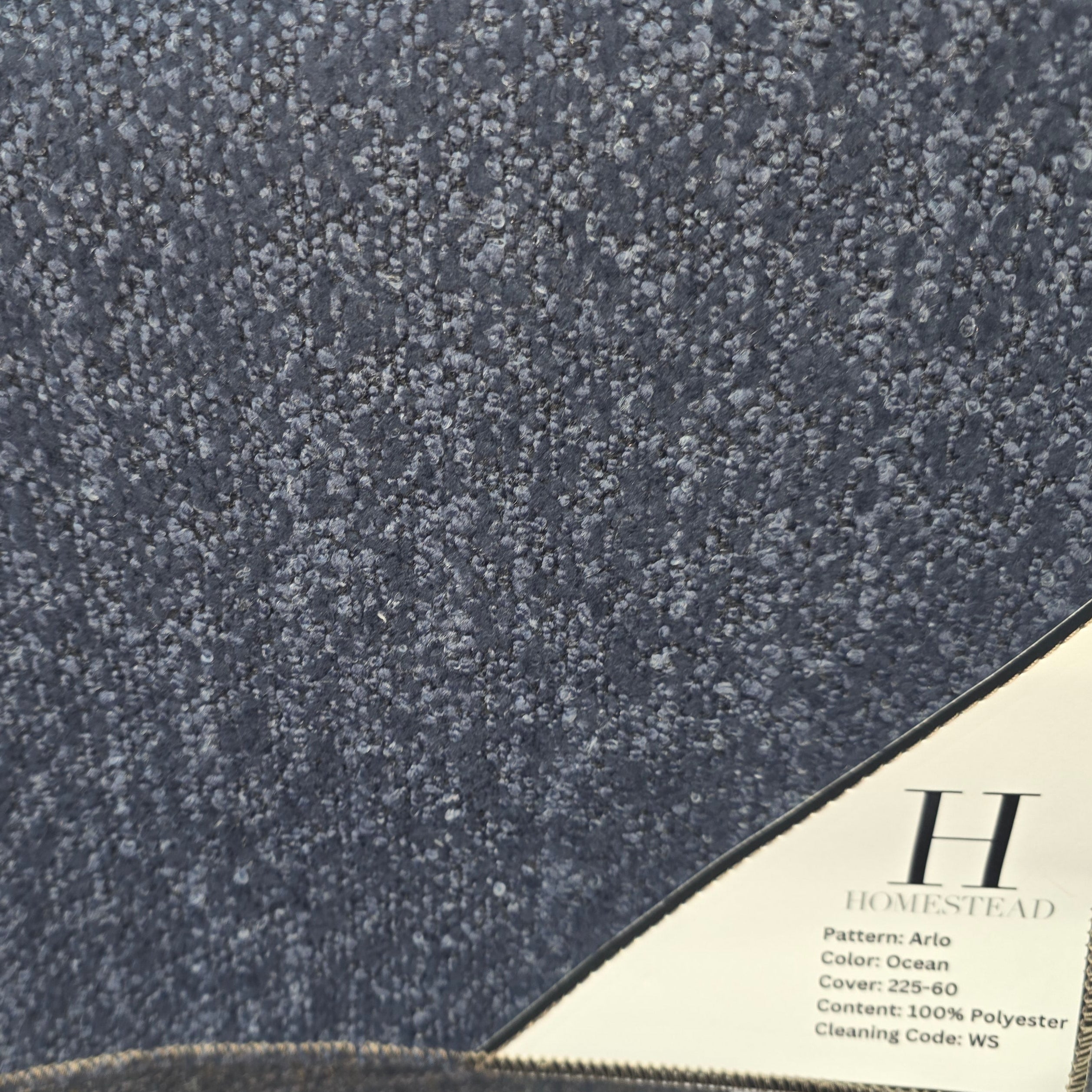 A close-up of a textured fabric or carpet sample in a dark heathered blue-gray tone, with a cream label attached in the lower right corner reading Homestead.