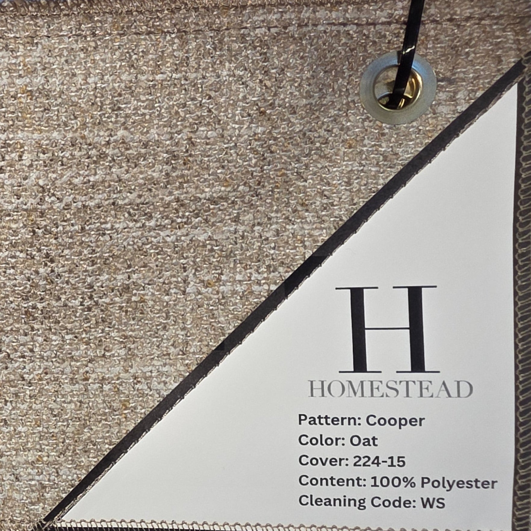 A close-up of a fabric sample card labeled H Homestead, showing a warm oatmeal-beige woven texture with a white printed label, hanging in front of other fabric swatches.