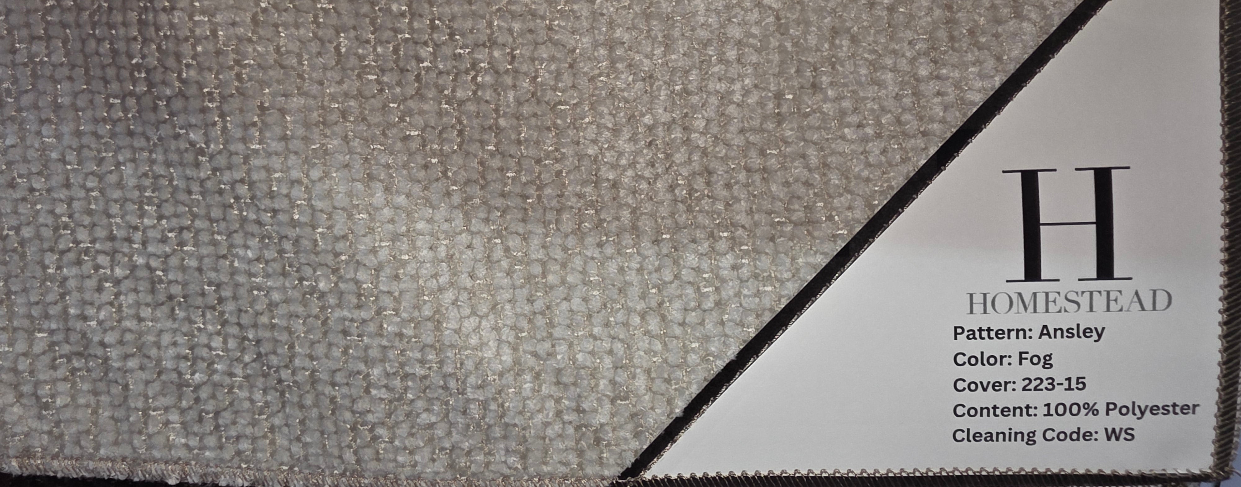 A close-up of a carpet sample swatch labeled Homestead Ansley, showing a textured light gray and beige patterned fabric with a white paper label attached.