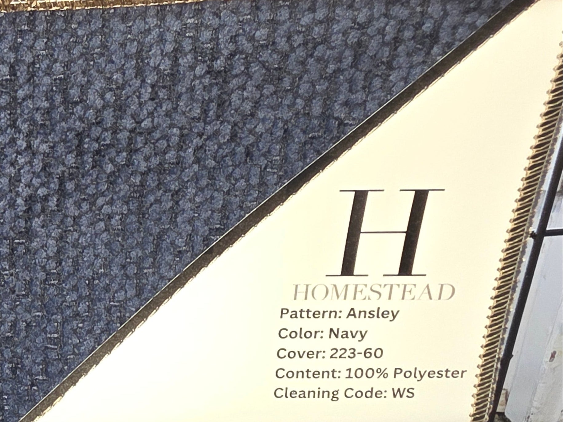 A close-up of a fabric or carpet sample card labeled 'H Homestead' with a navy textured woven area and a tan sample area above it, photographed in a showroom or office setting.