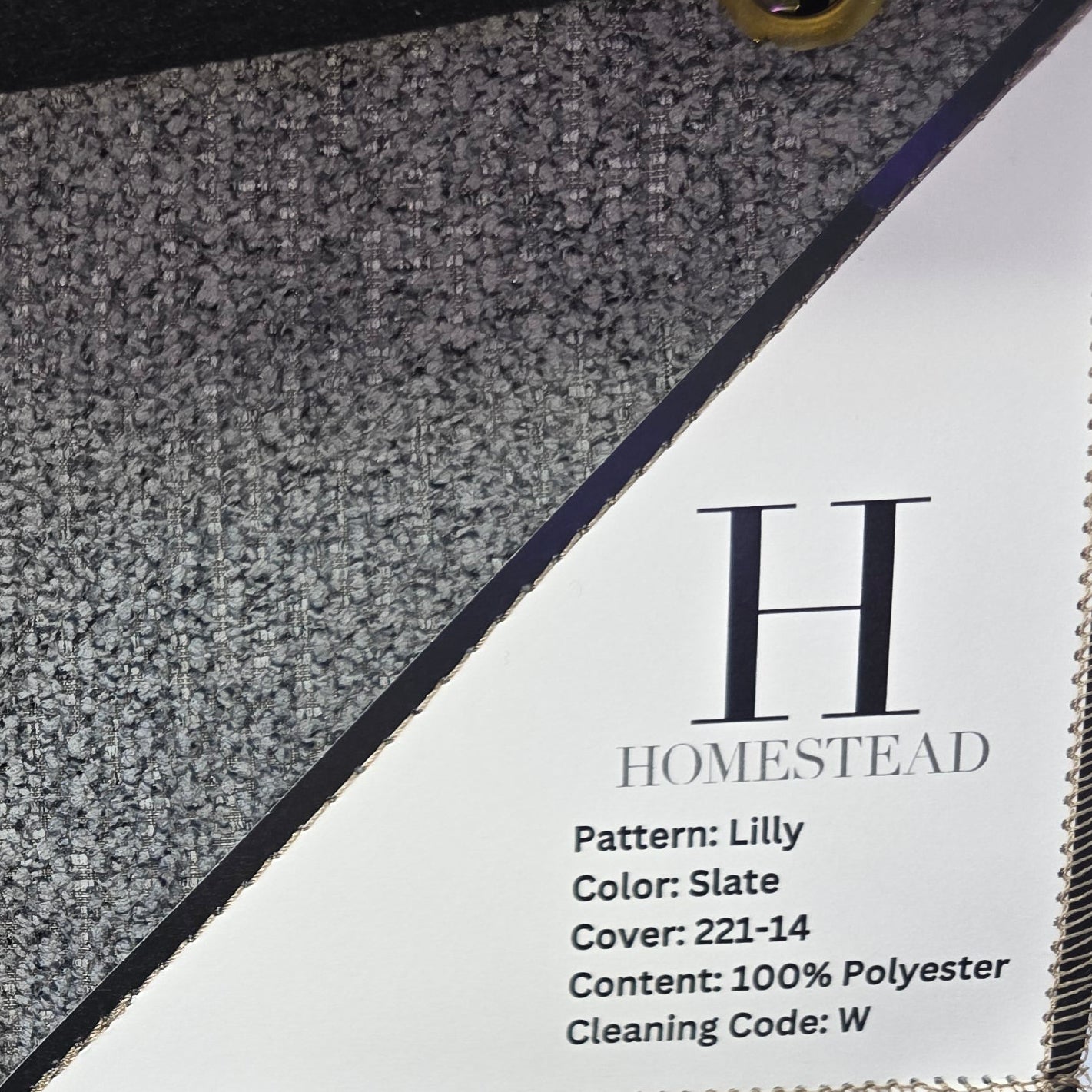 Two fabric or carpet swatches hanging on a display rack, each with a white label reading H Homestead and Pattern: Lilly, Color: Slate, Cover: 222-14, Content: 100% Polyester, Cleaning Code: W.