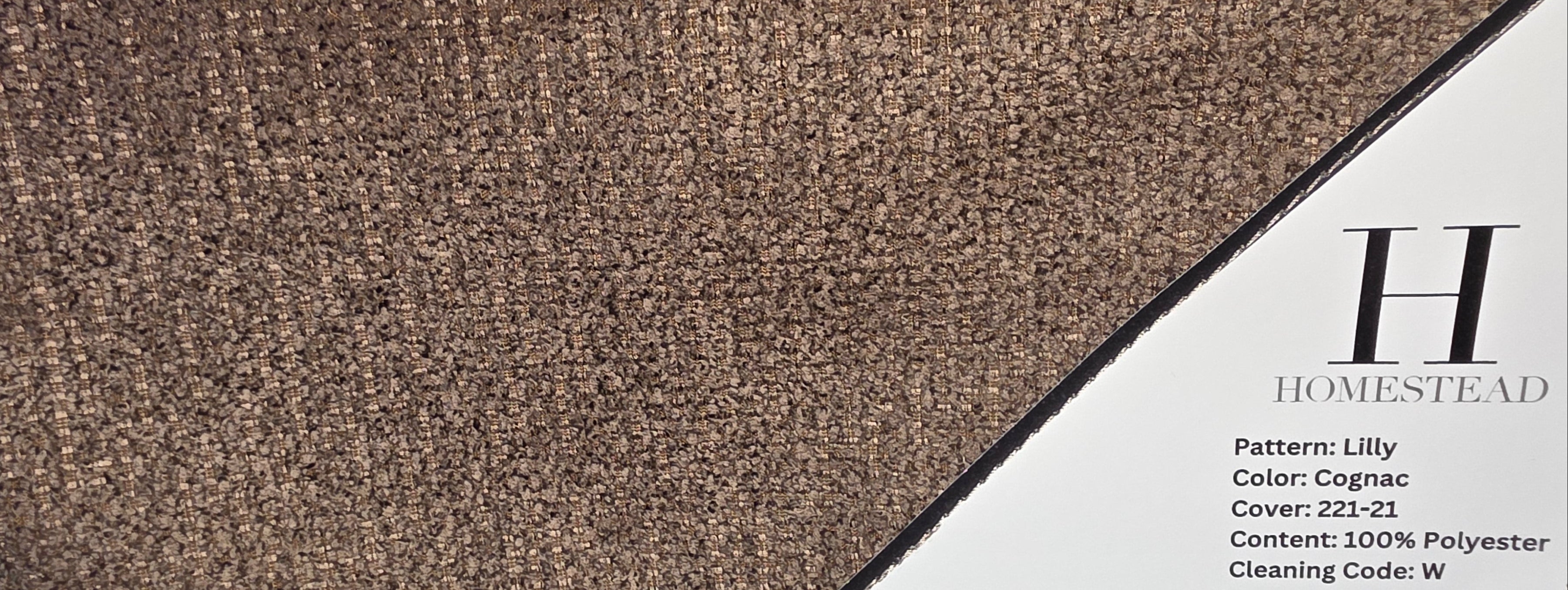 A carpet sample display showing a textured area rug in a warm taupe-brown color, with a printed product card labeled Homestead visible on the right side.