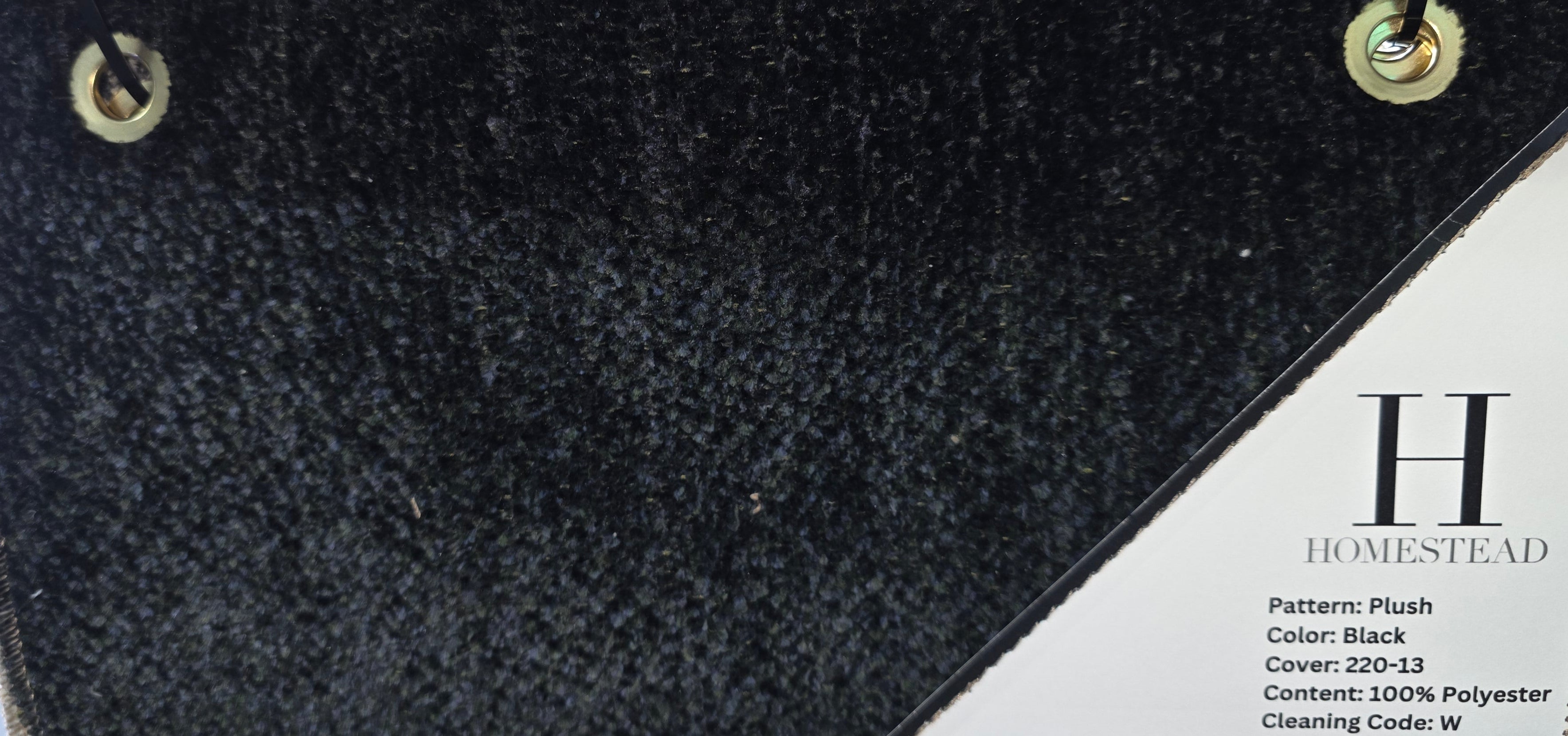 A close-up of a fabric or upholstery sample card showing a dark charcoal/black plush texture on the left and a light beige woven fabric on the right, with a printed label reading H Homestead and product details.