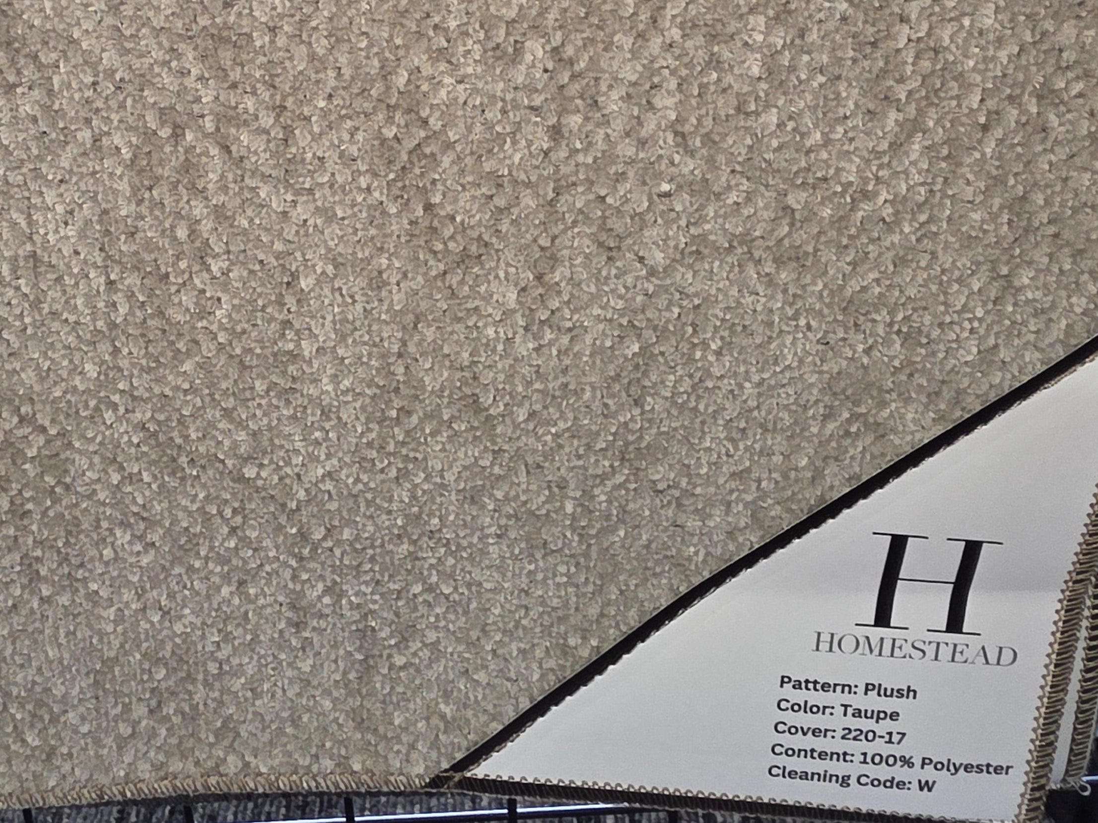 A large carpet sample swatch board from Homstead, showing a textured beige and taupe patterned pile, displayed on a black stand.