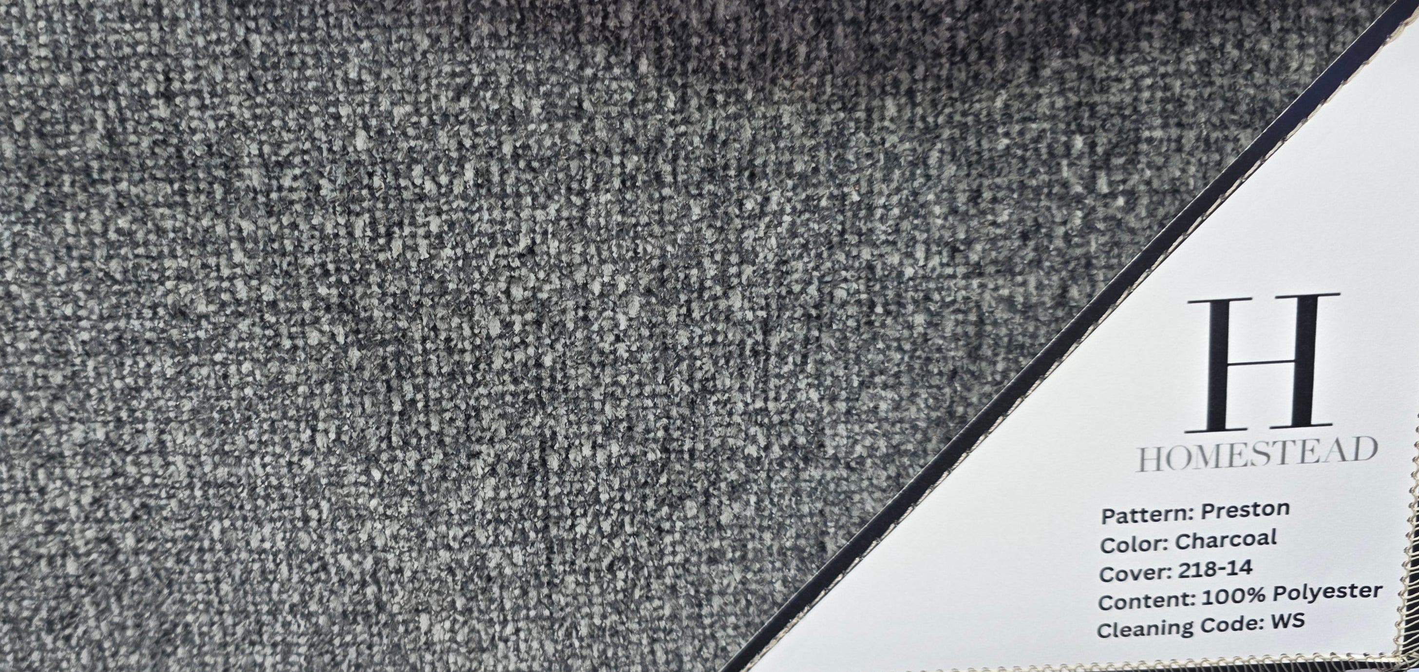 A close-up of a fabric sample card from Homestead, showing a charcoal gray woven textile with a white printed label. The card is hanging from metal grommets on a black wire rack.