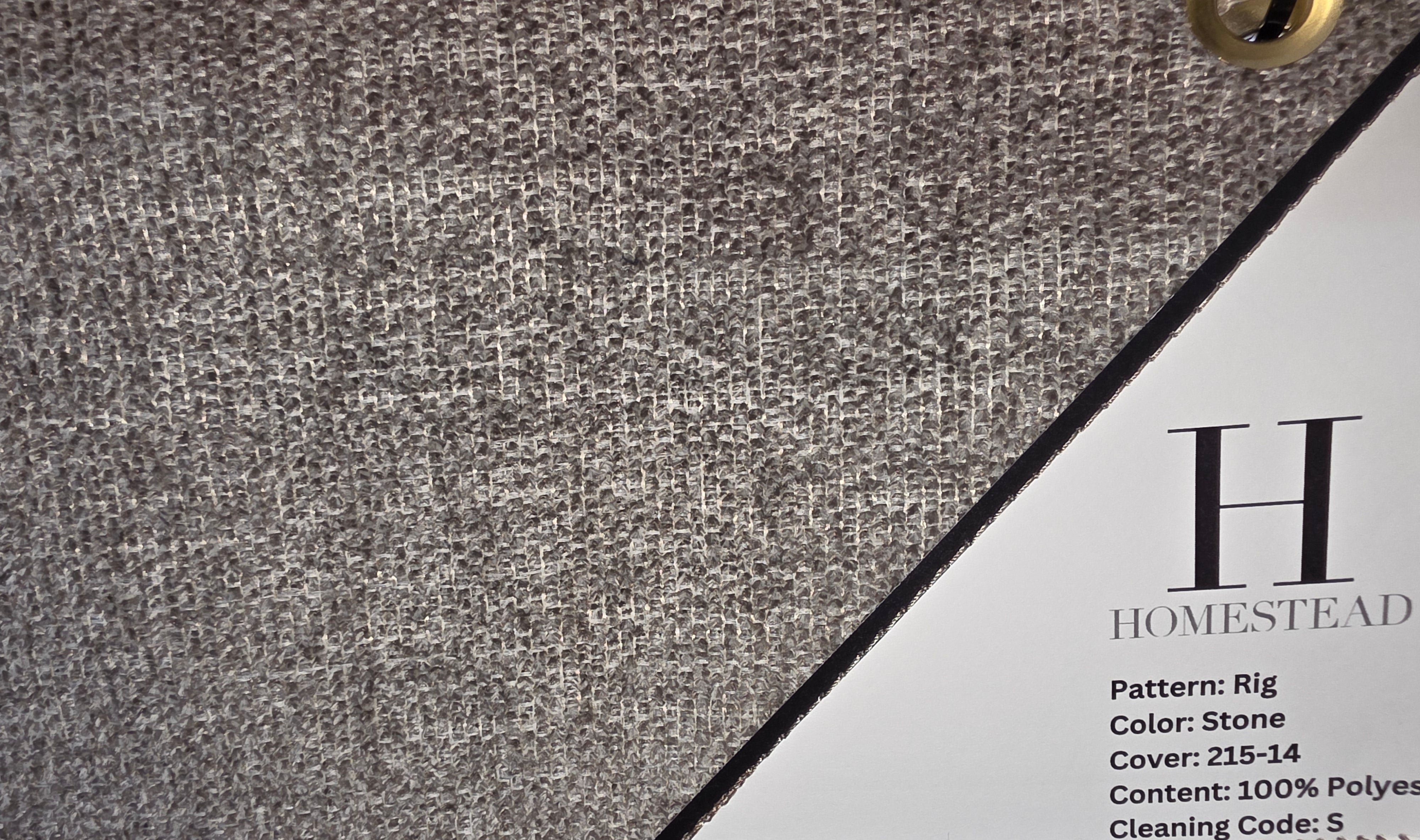 Close-up of a woven fabric swatch in a gray stone texture, with a white paper label corner visible at the bottom right showing the Homestead brand and pattern details.