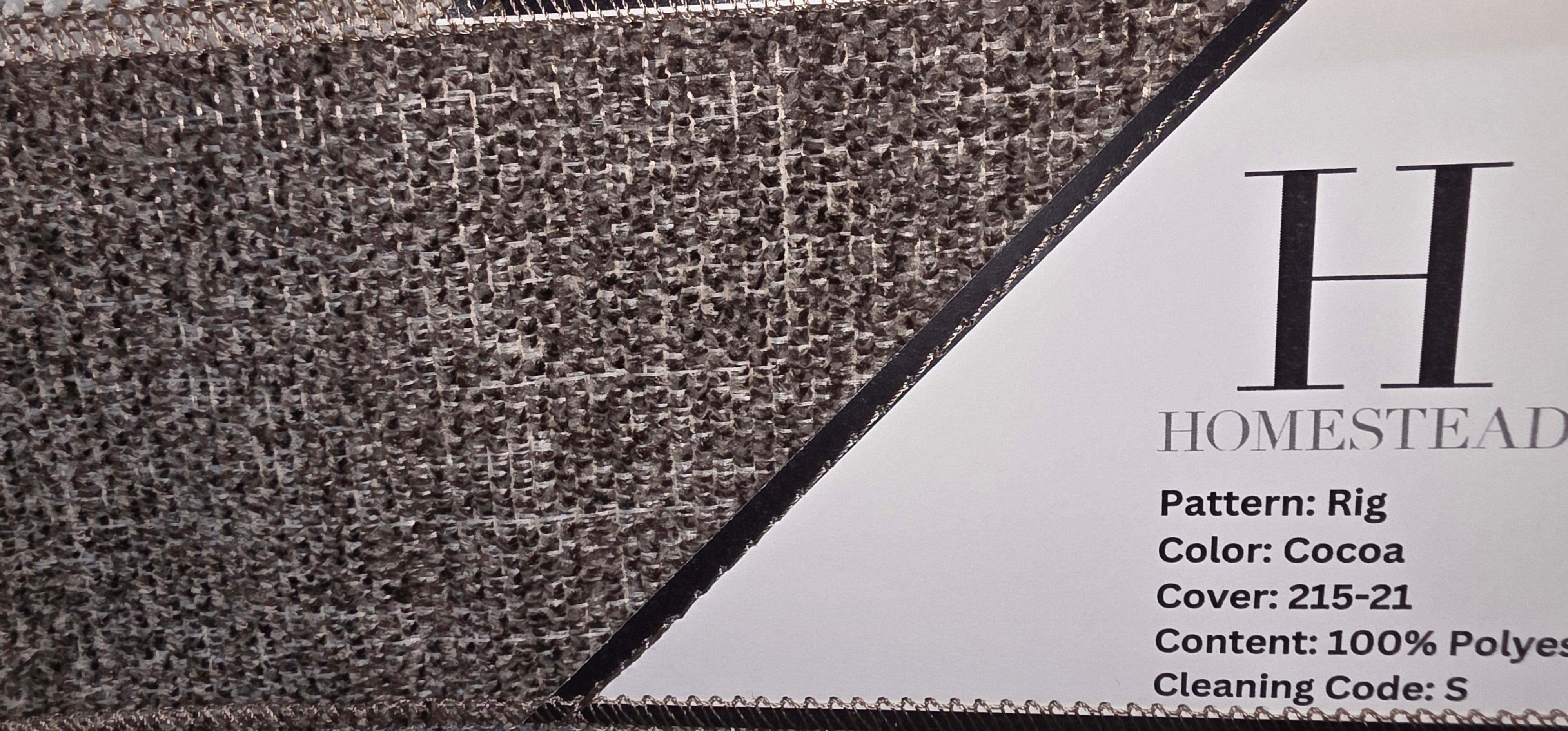 Close-up of a woven fabric swatch sample with a textured pattern, showing a white label with printed product details including 'HOMESTEAD' and color information.